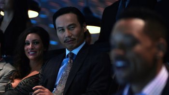 Movie still from “Focus” (2015), directed by John Requa – A man in a suit sitting in front of other people; Close Up shot, Over the shoulder angle