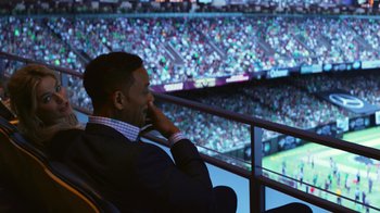 Movie still from “Focus” (2015), directed by John Requa – A man sitting in front of a large screen in a stadium; Medium shot, High angle