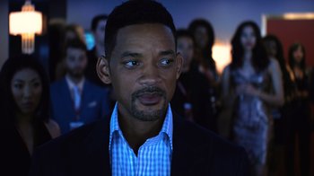 Movie still from “Focus” (2015), directed by John Requa – A man in a suit and tie in front of a group of people; Close Up shot, Over the shoulder angle