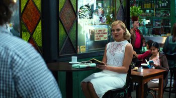 Movie still from “Focus” (2015), directed by John Requa – A woman sitting at a table in front of a store window; Medium shot, Over the shoulder angle