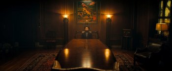Movie still from “Fool's Paradise” (2023), directed by Charlie Day – A man sitting at a large wooden table in front of a painting; Wide shot, High angle