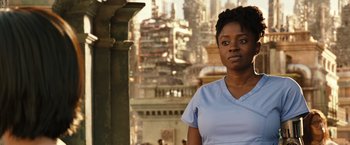 Movie still from “Alita: Battle Angel” (2019), directed by Robert Rodriguez – A woman standing in front of a building with buildings in the background; Close Up shot, Over the shoulder angle