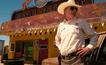 Movie still from “From Dusk Till Dawn” (1996), directed by Robert Rodriguez – A man in a cowboy hat and sunglasses standing in front of a building; Medium shot, Over the shoulder angle