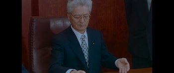 Movie still from “Fugitive: The Curious Case of Carlos Ghosn” (2022), directed by Lucy Blakstad – An older man sitting at a desk in a suit and tie; Medium shot, Low angle