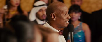 Movie still from “Furious 7” (2015), directed by James Wan – A man is speaking into a microphone in front of a group of people; Close Up shot, Over the shoulder angle