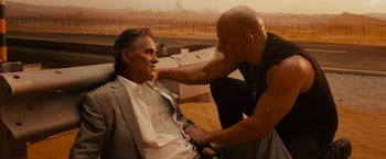 Movie still from “Furious 7” (2015), directed by James Wan – A man sitting next to another man on a beach; Medium shot, Over the shoulder angle