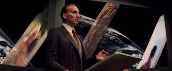 Movie still from “G.I. Joe: The Rise of Cobra” (2009), directed by Stephen Sommers – A man in a suit and tie standing in front of an image of the earth; Medium shot, Low angle