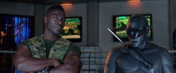 Movie still from “G.I. Joe: The Rise of Cobra” (2009), directed by Stephen Sommers – A man standing next to another man in front of a television; Medium shot, Over the shoulder angle