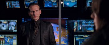 Movie still from “G.I. Joe: The Rise of Cobra” (2009), directed by Stephen Sommers – A man in a suit and tie standing in front of a television; Medium shot, Over the shoulder angle