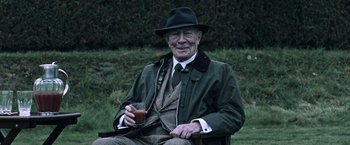 Movie still from “All the Money in the World” (2017), directed by Ridley Scott – An older man in a hat and a jacket holding a drink; Medium shot, High angle
