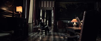 Movie still from “All the Money in the World” (2017), directed by Ridley Scott – A man sitting in a chair in a room with black and white checkered floor; Wide shot, Low angle