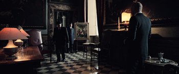 Movie still from “All the Money in the World” (2017), directed by Ridley Scott – Two men in suits and hats in a room with a painting; Wide shot, Low angle