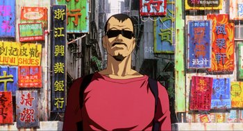 Movie still from “Ghost in the Shell” (1995), directed by Mamoru Oshii – A man in a red shirt is standing in front of some buildings; Medium shot, Low angle