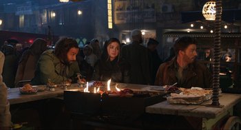 Movie still from “Ghosted” (2023), directed by Dexter Fletcher – A group of people sitting around a fire pit; Medium shot, Over the shoulder angle