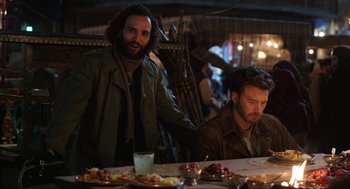 Movie still from “Ghosted” (2023), directed by Dexter Fletcher – Two men sitting at a table with plates of food in front of them; Medium shot, Over the shoulder angle