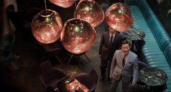 Movie still from “Ghosted” (2023), directed by Dexter Fletcher – Two men standing in front of a table and chairs; Wide shot, Overhead angle