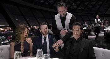 Movie still from “Ghosted” (2023), directed by Dexter Fletcher – A group of people sitting at a table with a man cutting another man's hair; Medium shot, Low angle