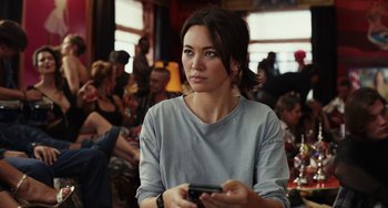 Movie still from “Glass Onion” (2022), directed by Rian Johnson – A woman holding a cell phone while standing in front of a group of people; Close Up shot, Over the shoulder angle