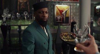 Movie still from “Glass Onion” (2022), directed by Rian Johnson – A man in a green suit standing next to a table; Close Up shot, Over the shoulder angle