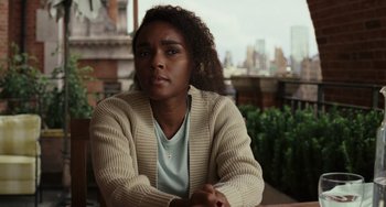 Movie still from “Glass Onion” (2022), directed by Rian Johnson – A woman sitting at a table in front of a city skyline; Close Up shot, Over the shoulder angle