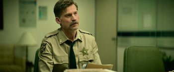 Movie still from “God Is a Bullet” (2023), directed by Nick Cassavetes – A man in a sheriff's uniform sitting at a table; Medium shot, Over the shoulder angle