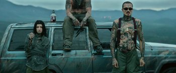 Movie still from “God Is a Bullet” (2023), directed by Nick Cassavetes – A man sitting on the hood of a car with a pair of scissors; Medium shot, Low angle