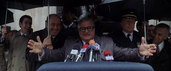 Movie still from “Godzilla” (1998), directed by Roland Emmerich – A man in a gray suit is giving a press conference with microphones in front of him; Medium shot, Low angle