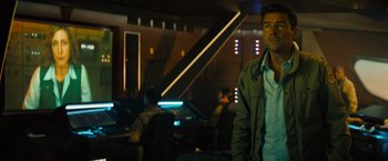 Movie still from “Godzilla: King of the Monsters” (2019), directed by Michael Dougherty – A man standing in front of a group of people in a room; Medium shot, Over the shoulder angle