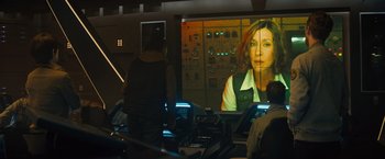 Movie still from “Godzilla: King of the Monsters” (2019), directed by Michael Dougherty – A woman is on a video screen in a room; Medium shot, Over the shoulder angle