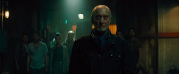 Movie still from “Godzilla: King of the Monsters” (2019), directed by Michael Dougherty – An older man standing in a dark room with a crowd; Medium shot, Low angle