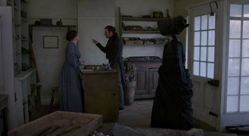 Movie still from “Ammonite” (2020), directed by Francis Lee – Two women and a man dressed in period clothing in an old - fashioned kitchen; Medium shot, Over the shoulder angle