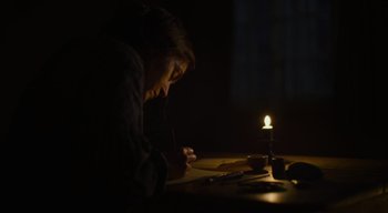 Movie still from “Ammonite” (2020), directed by Francis Lee – A person sitting at a table with a candle in a dark room; Medium shot, Low angle