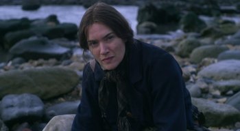 Movie still from “Ammonite” (2020), directed by Francis Lee – A woman sitting on a rock near a body of water; Close Up shot, High angle