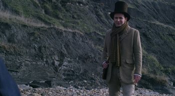 Movie still from “Ammonite” (2020), directed by Francis Lee – A man wearing a hat and holding a gun standing on a rocky beach; Medium shot, Low angle