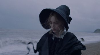 Movie still from “Ammonite” (2020), directed by Francis Lee – A young woman wearing a black hat standing on the beach; Close Up shot, High angle