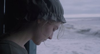 Movie still from “Ammonite” (2020), directed by Francis Lee – A young woman wearing a hat looking out of a window; Close Up shot, Over the shoulder angle