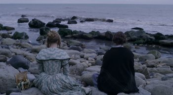Movie still from “Ammonite” (2020), directed by Francis Lee – Two people sitting on rocks near the water; Wide shot, High angle