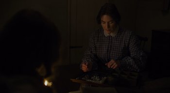 Movie still from “Ammonite” (2020), directed by Francis Lee – A woman sitting at a table in a dimly - lit room; Medium shot, High angle
