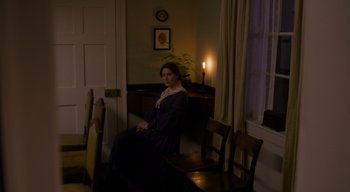 Movie still from “Ammonite” (2020), directed by Francis Lee – A woman sitting at a table in a room with a candle; Wide shot, High angle