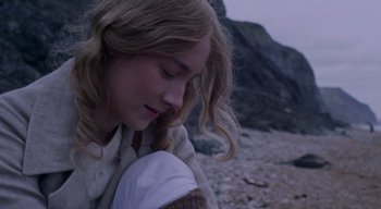 Movie still from “Ammonite” (2020), directed by Francis Lee – A young girl sitting on the beach looking down at the ground; Close Up shot, High angle