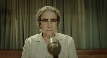 Movie still from “Golda” (2023), directed by Guy Nattiv – An older woman wearing glasses is sitting in front of a microphone; Close Up shot, High angle