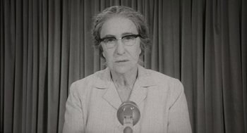 Movie still from “Golda” (2023), directed by Guy Nattiv – An older woman wearing glasses is speaking into a microphone in front of a curtain; Close Up shot, High angle