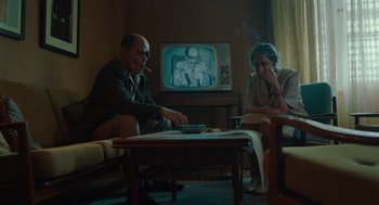 Movie still from “Golda” (2023), directed by Guy Nattiv – A man and a woman sitting in front of a television; Medium shot, Over the shoulder angle