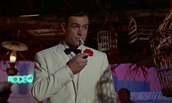 Movie still from “Goldfinger” (1964), directed by Guy Hamilton – A man in a white suit smoking a cigarette and holding a red flower; Medium shot, Low angle