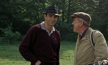 Movie still from “Goldfinger” (1964), directed by Guy Hamilton – A man wearing a hat talking to another man in a field; Medium shot, Over the shoulder angle
