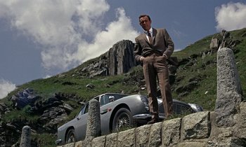 Movie still from “Goldfinger” (1964), directed by Guy Hamilton – A man standing next to a car on top of a hill; Wide shot, Low angle