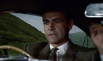 Movie still from “Goldfinger” (1964), directed by Guy Hamilton – A man wearing a suit and tie driving a car; Close Up shot, Low angle