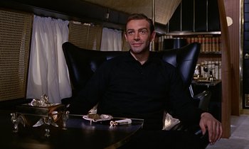 Movie still from “Goldfinger” (1964), directed by Guy Hamilton – A man sitting at a table in a black chair; Medium shot, Low angle