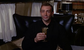 Movie still from “Goldfinger” (1964), directed by Guy Hamilton – A man sitting in a chair holding a glass of wine; Medium shot, Low angle