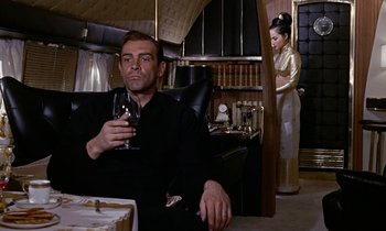 Movie still from “Goldfinger” (1964), directed by Guy Hamilton – A man and a woman sitting in a room drinking wine; Medium shot, Low angle